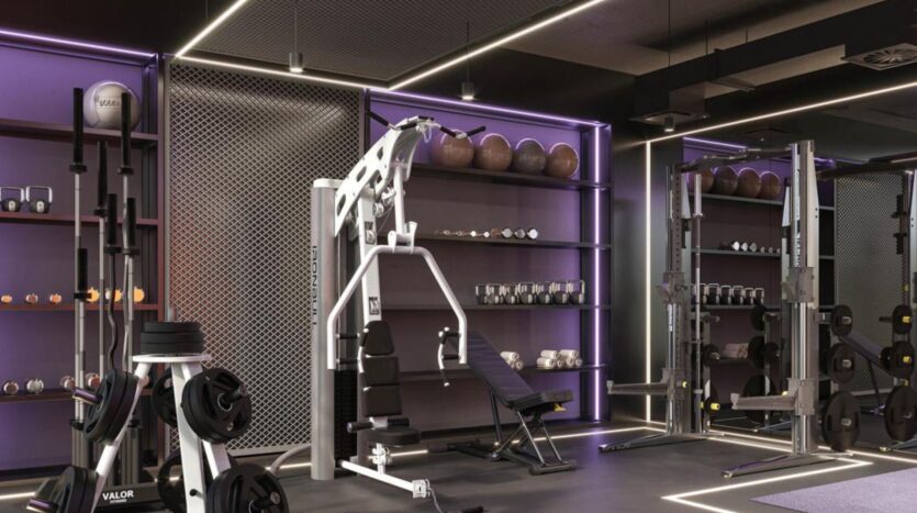 Modern gym in Rabdan Gardens with a variety of equipment, including weight machines, free weights, medicine balls and benches. Purple accent lighting highlights shelving and workout areas. The space is clean and organized.