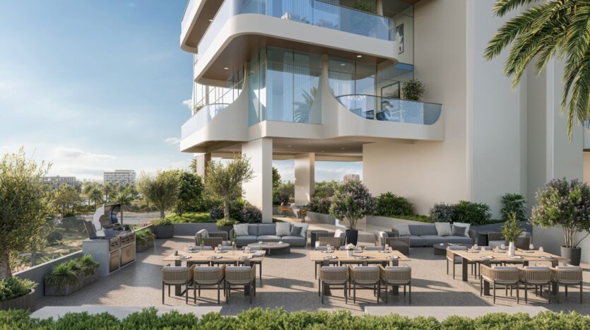 Sala da pranzo all'aperto con diversi tavoli e sedie disposti su un patio circondato dal verde. Lo spazio è sovrastato dal Caden, un edificio moderno con balconi in vetro e architettura curvilinea sotto un cielo limpido.