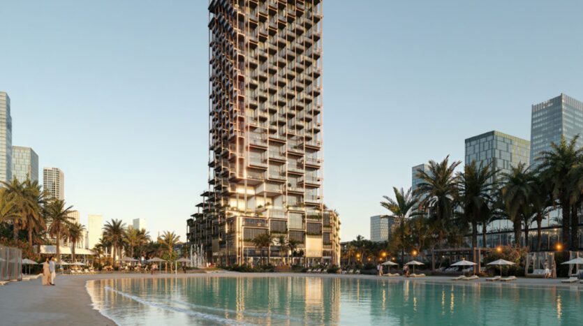 The Symphony, a modern skyscraper with a striking geometric facade, stands on the edge of a sandy beach lined with palm trees and calm turquoise water, surrounded by other contemporary skyscrapers under a clear sky.