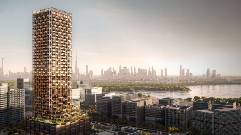 A large modern skyscraper with a unique grid façade, called The Symphony, stands among low-rise buildings, overlooking a river and a distant city under a cloudy sky.