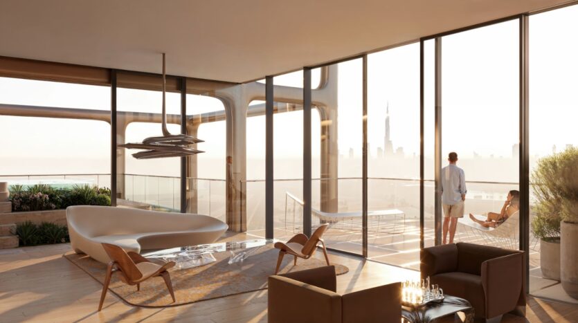 The Symphony's modern living room features floor-to-ceiling windows overlooking the city and balcony. A man stands near the railing while a woman relaxes in a hanging chair; elegant furniture and natural light fill the airy space.