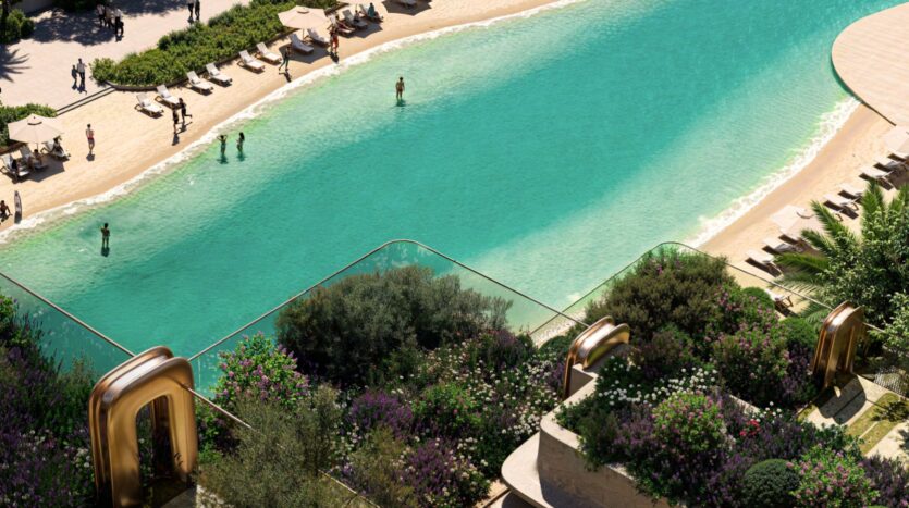 A rooftop garden with lush vegetation at Symphony overlooks a large turquoise pool, where people relax and swim near the sandy shoreline lined with sun loungers and umbrellas.