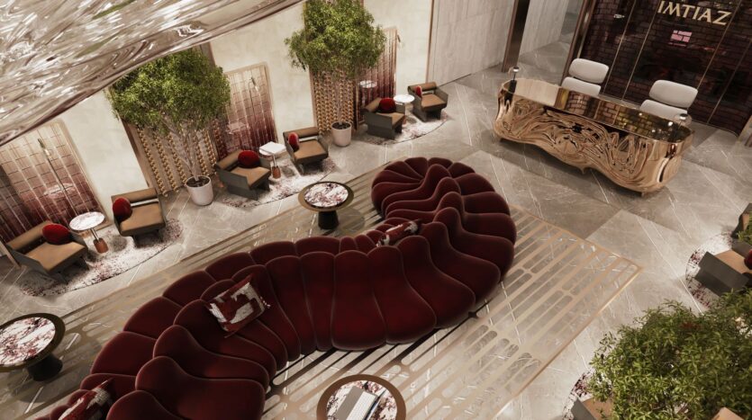 The lobby of a modern hotel, The Symphony, features a large burgundy velvet curved sofa, round tables, armchairs, large potted plants and a decorative gold reception desk. Gleaming marble floors and abundant light complete this sophisticated space.