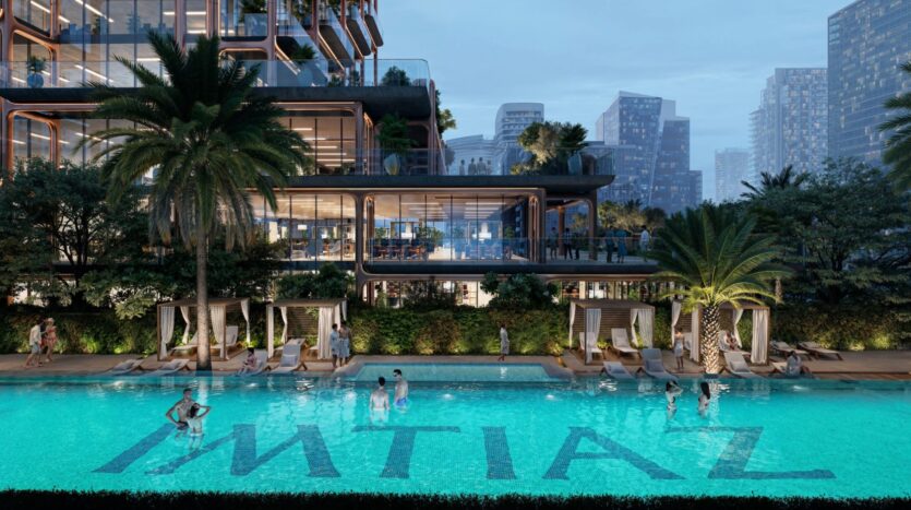 A modern, luxurious pool with "IMTIAZ" written on the floor, surrounded by palm trees, cabanas, and people relaxing. Behind it, the elegant, multi-story glass building of the Symphony rises, standing out against the city skyline at dusk.