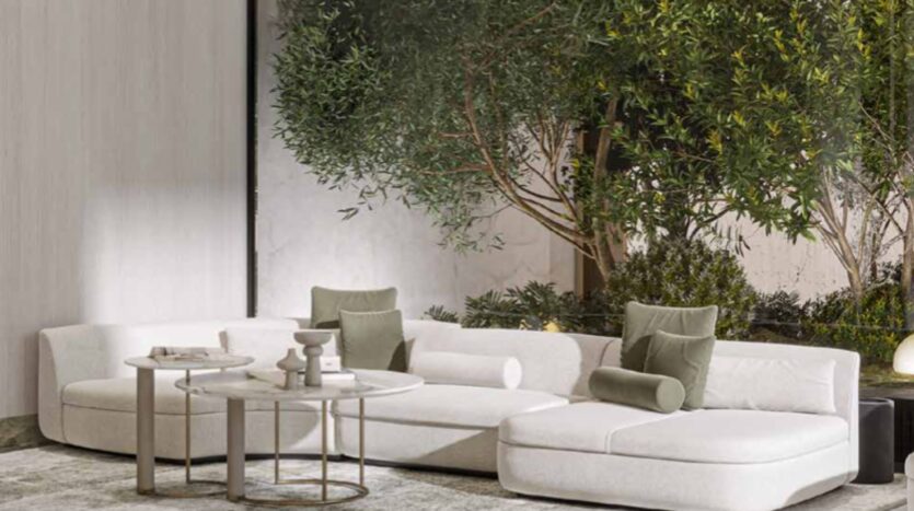 A modern living room with a white sectional sofa, green accent pillows, and nested round coffee tables, positioned next to large windows overlooking the trees and greenery of Arlington Park 2 outside.