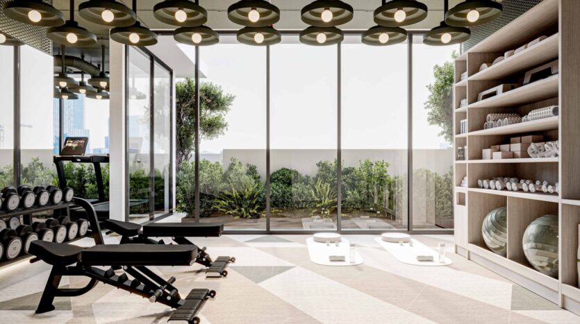 Modern gym at Arlington Park 2 with large windows, greenery outside, dumbbells, benches, a treadmill, yoga mats with towels, and shelves containing towels and exercise balls. Circular ceiling lights illuminate the bright, clean space.