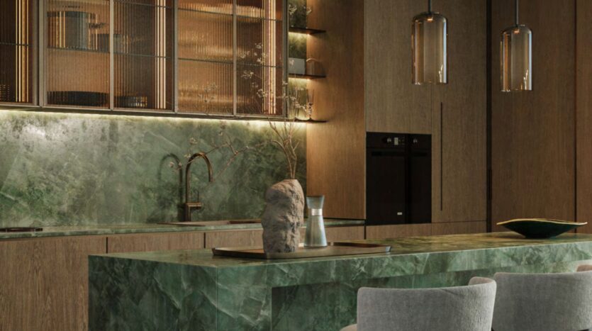 Modern kitchen with an island and green marble backsplash, wooden cabinets, open shelves with decor, built-in ovens, pendant lights, and two gray upholstered chairs. A tray with a vase and a Hado rock is placed on the island.
