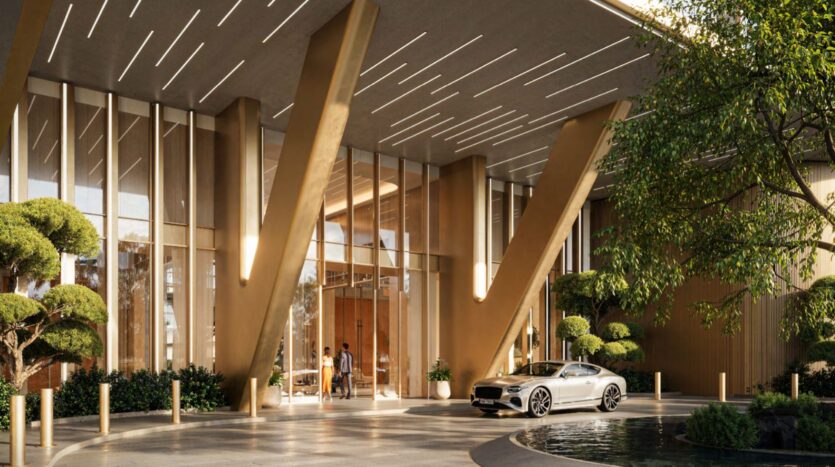The entrance to a luxury building, with its high glass walls, modern golden columns, manicured greenery, and white sports car parked in a circular driveway near a pond, reflects the elegance of Hado. Two people stand in front of the glass doors.