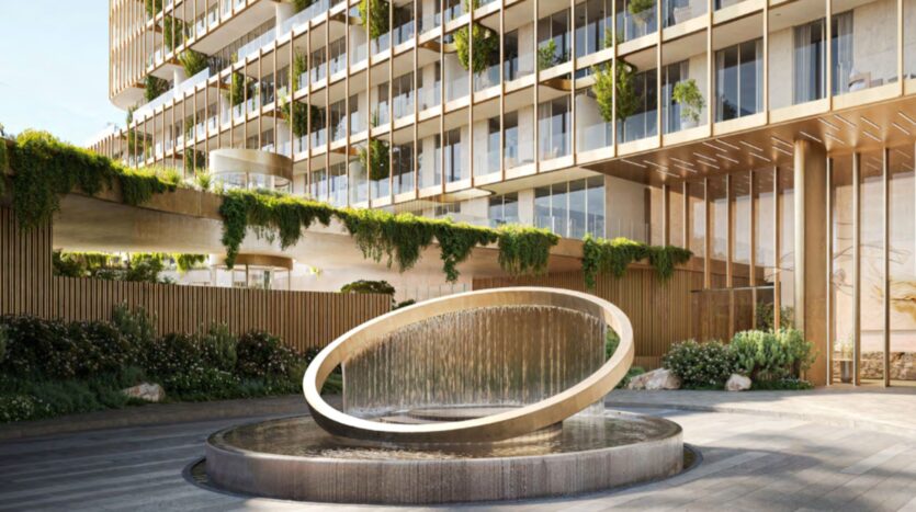 A modern Hado building with glass balconies and green plants, featuring a circular fountain with water cascading from a sloped ring in the landscaped entrance area.