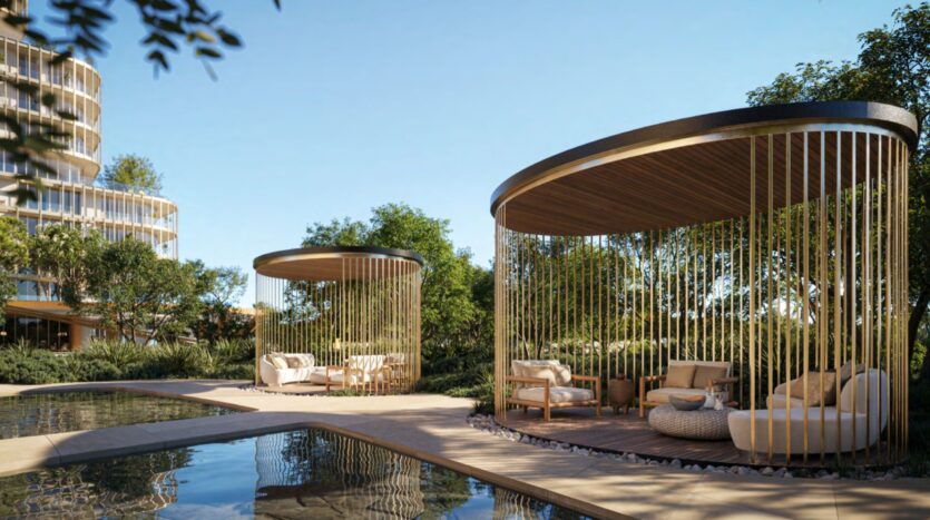 Two modern Hado outdoor pavilions, featuring wooden slats and comfortable seating, are located next to a reflective pool, surrounded by lush vegetation and trees under a clear blue sky.