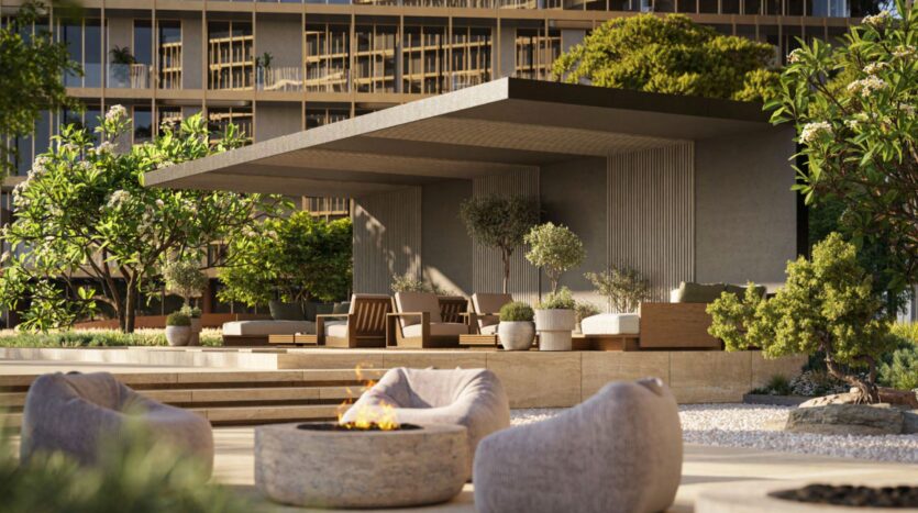 A modern outdoor lounge designed by Hado features upholstered seating under a shaded pavilion, surrounded by lush greenery, with a fireplace and rounded stone seating in the foreground. A residential building rises in the background.