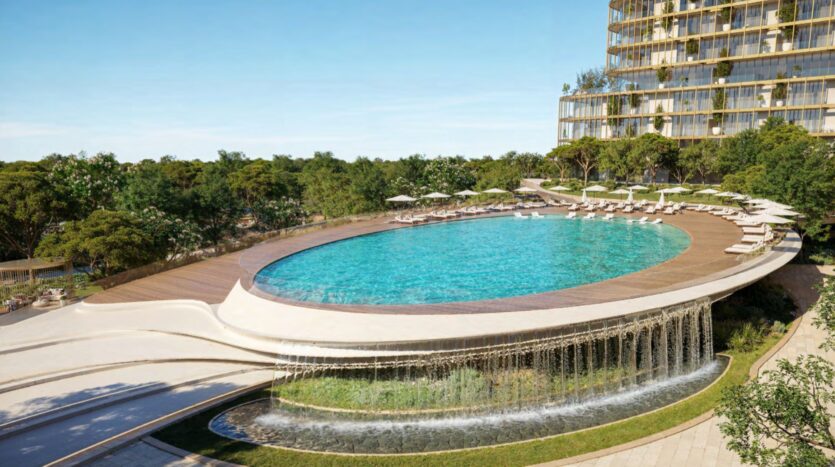 An oval-shaped Hado pool with a wooden deck, lounge chairs, and umbrellas. Water cascades over the edge of the pool. Trees surround the area, and a modern glass building stands to the right.