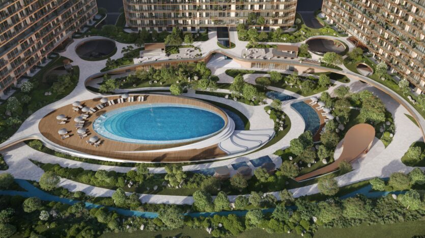 Aerial view of the courtyard of the modern Hado apartment complex, featuring an oval swimming pool, a wooden deck with lounge chairs, lush greenery, winding paths, and a tranquil stream.