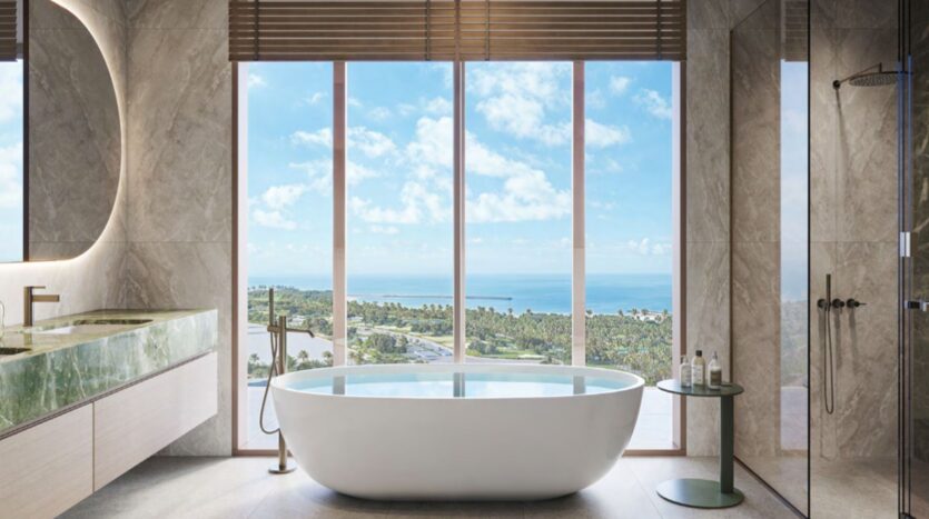 A modern Hado bathroom with a freestanding white bathtub faces large floor-to-ceiling windows, offering picturesque views of the greenery and the ocean. The room features a glass shower, marble walls, and an elegant vanity unit.