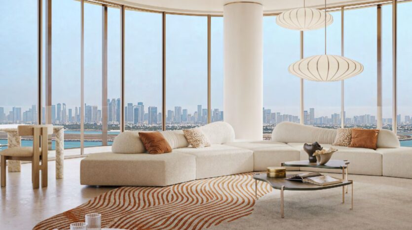 A modern living room with cream-colored sofas, a glass coffee table, and a decorative rug features Hado-inspired accents. Floor-to-ceiling windows overlook the city skyline and waterfront, while pendant lights add a contemporary touch.