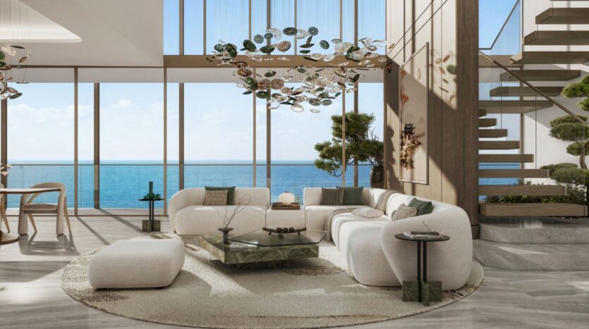 Modern living room with large windows overlooking the ocean, light-colored furniture, a round Hado-inspired rug, a glass coffee table, indoor plants, contemporary chandeliers, and a staircase on the right side.