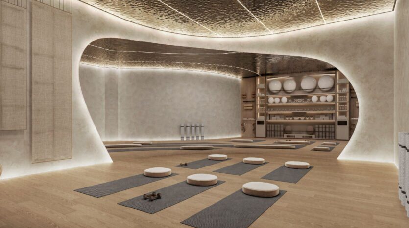 A modern, minimalist yoga studio with wooden floors, soft lighting, and round cushions on black mats. The curved walls and ceiling evoke the calming energy of Hado, while neat shelves hold yoga accessories along the back wall.