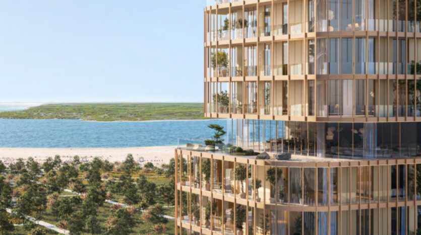 A modern, high-rise Hado-style building with numerous bay windows and balconies adorned with greenery, overlooking lush scenery and a stretch of water under a clear blue sky.