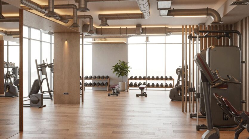 The Holm One gym is spacious and modern, with wooden floors, large windows, weights and dumbbells, exercise machines, and a potted plant in the center, all bathed in natural light.