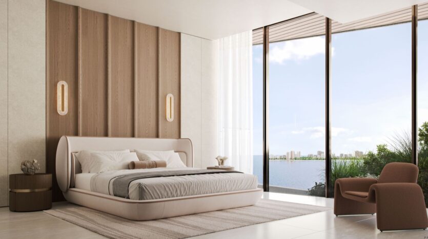 Modern bedroom with a large bed, touches of beige and wood, a side table, an armchair, and floor-to-ceiling windows offering Lia a breathtaking view of the water and the city skyline. Natural light fills the minimalist and serene space.