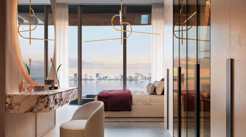 Lia's modern bedroom with large windows overlooking the city and water, featuring a marble vanity, comfortable chair, bed with red sheets, mirrored closet doors, and elegant gold pendant lights.