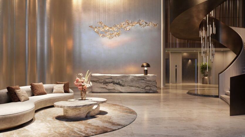 The modern lobby of the Lia Hotel features a curved beige sofa, marble coffee tables, a sculptural reception desk, hanging artwork, and a dramatic spiral staircase with cascading lights. The space highlights neutral tones and elegant décor.