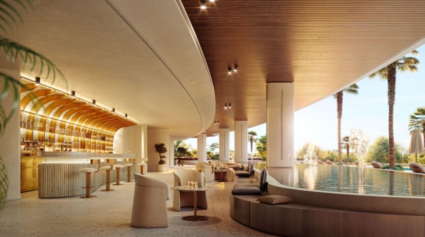 This modern and luxurious hotel lounge features a curved wooden ceiling, an elegant bar, plush seating, and a round indoor pool. Large windows frame the palm trees and bright outdoor landscape, creating a welcoming retreat at Lia.