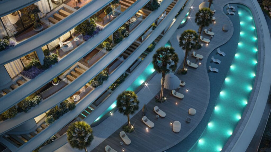 Aerial view of Lia's modern, curved building, whose balconies overlook a pool lit by blue lights. Palm trees, lounge chairs, and small tables are arranged on a wooden deck by the pool as night falls.
