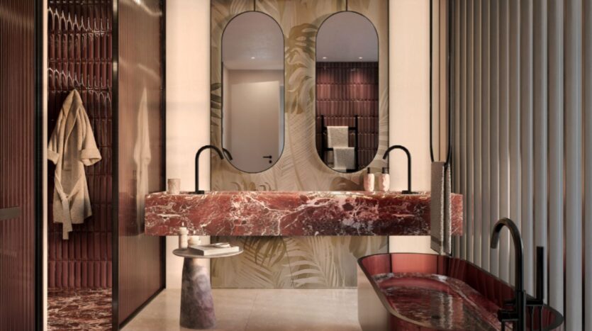 A modern Lia bathroom with a double red marble sink, two curved mirrors, a matching red marble bathtub, a hanging bathrobe, and textured wall tiles in warm tones, accentuated by soft lighting.