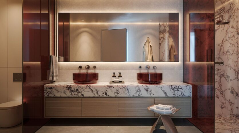 Modern bathroom with a marble countertop, two red glass sinks, and red accents inspired by Lia. A large backlit mirror and a wooden stool with towels enhance the luxurious and contemporary look of the elegant marble and wood textures.