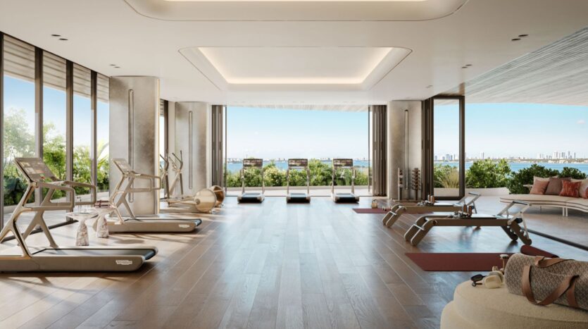 The Lia gym is spacious and modern, with large windows overlooking the city skyline. It is equipped with treadmills, Pilates reformers, yoga mats, and fitness equipment on light wood flooring, surrounded by greenery outside.