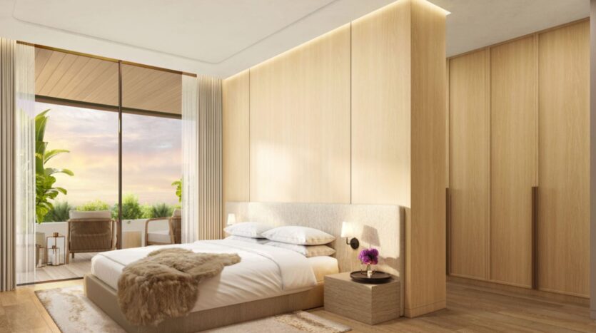 The modern, minimalist bedroom at The Edition features a large bed, light wood paneling, nightstands, and floor-to-ceiling windows that open onto a balcony where you can sit outdoors and admire the lush greenery at sunset.