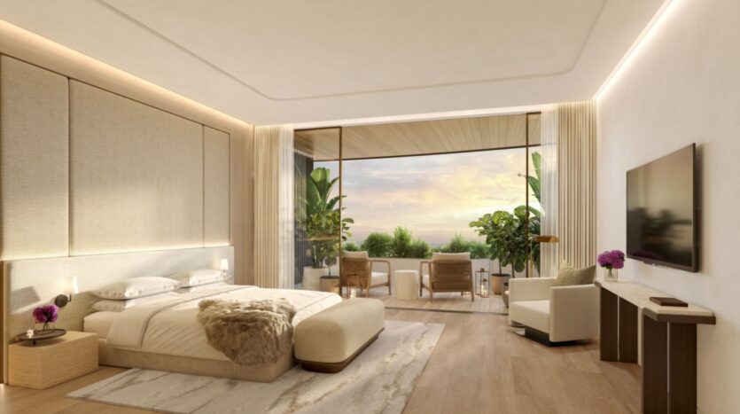 Elegant and modern room at The Edition hotel, featuring a large bed, soft lighting, a wall-mounted television, and a sitting area. Floor-to-ceiling windows open onto a terrace with chairs, lush plants, and breathtaking views of the sunset.