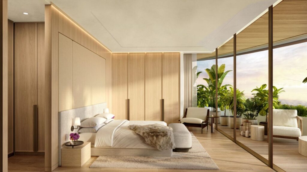 The modern bedroom at The Edition hotel features light wood walls and floors, a large bed, bedside tables, a comfortable armchair, and floor-to-ceiling windows that open onto a lush balcony decorated with green plants and outdoor seating. Natural light fills the room.