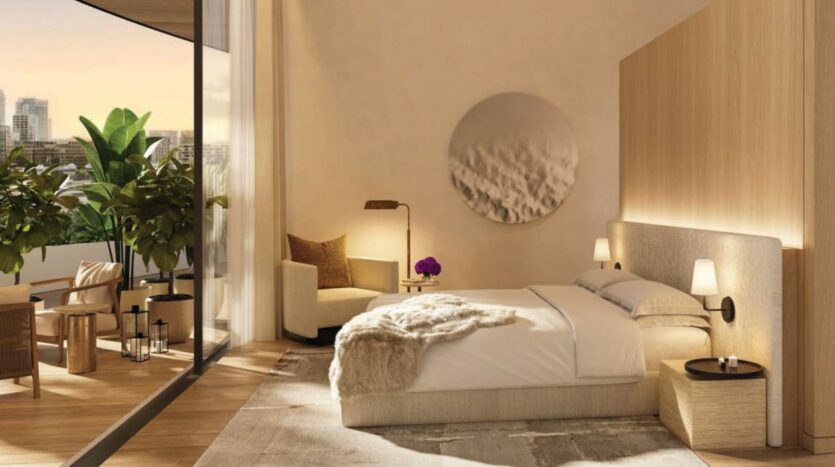 A modern room at The Edition with a large bed, neutral décor, and soft lighting. Floor-to-ceiling windows open onto a balcony offering views of the city and lush plants. A comfortable armchair, side tables, and wall art complete this serene space.