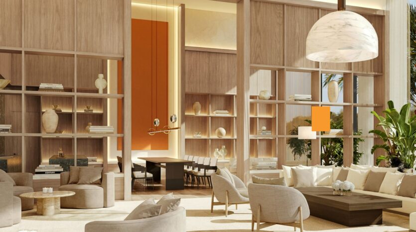 Modern, sunlit living room from the Edition with cream-colored sofas, round tables, and open wooden shelves displaying vases and books. A dining room is visible in the background, separated by tall shelves. A large pendant lamp hangs above.