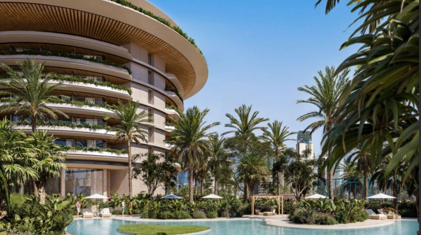 The luxurious curved building of the Edition, featuring balconies, overlooks a tropical pool surrounded by palm trees, lush vegetation, and sun loungers shaded by white umbrellas on sunny days.