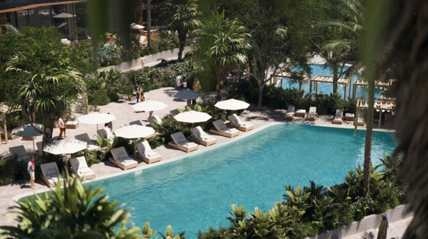 A lush outdoor pool at The Edition, surrounded by tropical plants, with lounge chairs and white umbrellas by the pool. People relax and stroll around on a clear, sunny day.