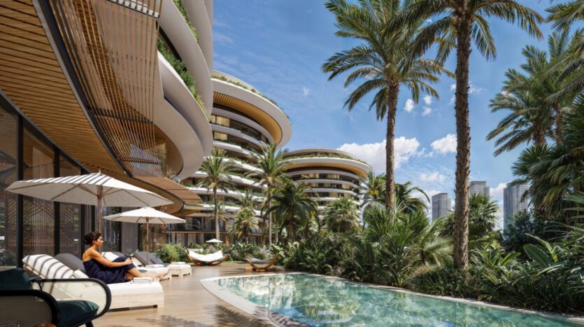 A woman relaxes by the pool at The Edition, surrounded by palm trees and modern, curved residential buildings, with the city's skyscrapers visible in the background under a bright blue sky.
