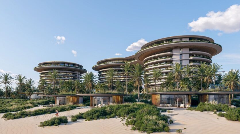 The modern, circular buildings of The Edition waterfront, with large balconies and green roofs, rise behind palm trees and sand dunes, under a blue sky with scattered clouds. A person walks on the sand in the foreground.