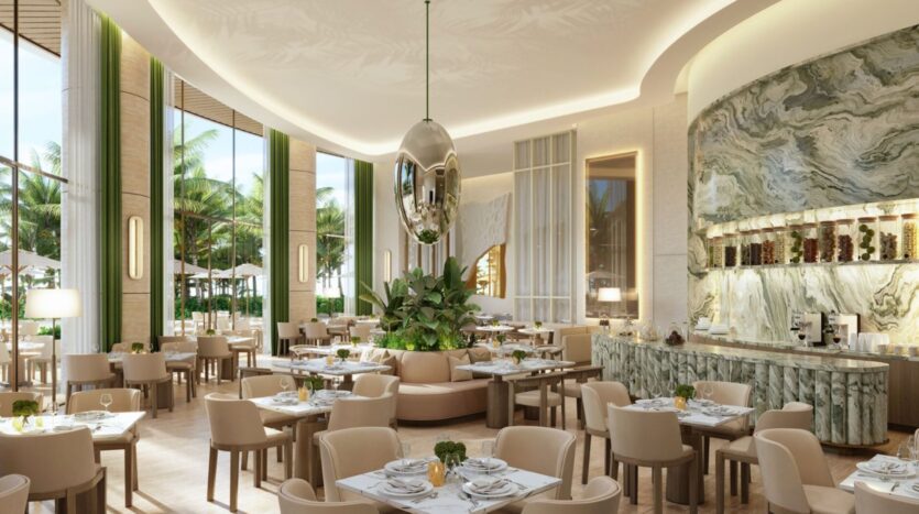 This modern and elegant restaurant at The Edition features beige chairs and tables, a central plant arrangement, large windows overlooking the palm trees outside, and a marble accent wall with shelves and a coffee station. Soft lighting creates a welcoming atmosphere.