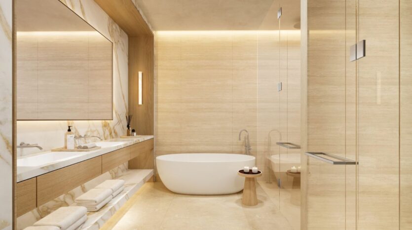 The modern bathroom in the Edition features beige stone walls, a freestanding white bathtub, a wooden stool with candles, a double sink with a large mirror, and towels neatly stacked on open shelves beneath the counter.