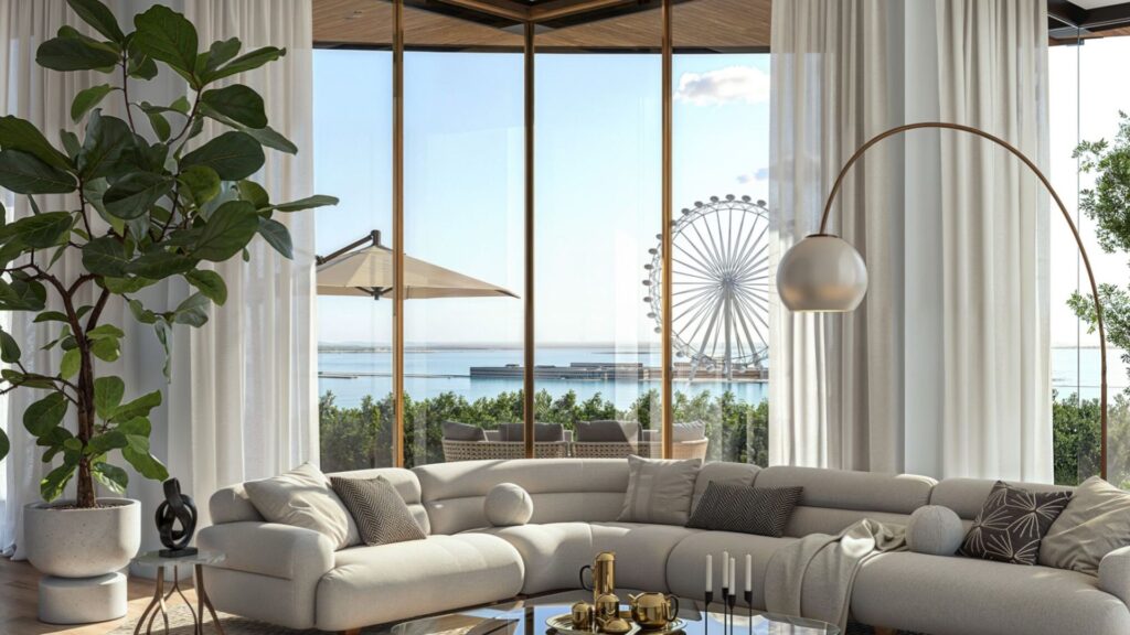 The modern living room at the Edition features a large curved beige sofa, a glass coffee table, lush plants, and tall windows offering views of the Ferris wheel, the ocean, and a patio with a parasol. Light floods this airy and elegant space.