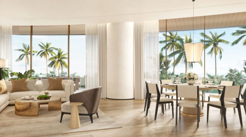 The modern, open-plan living and dining room at The Edition hotel features neutral décor, large windows with views of palm trees and blue skies, a round dining table, a comfortable sofa, an armchair, light wood floors, and elegant lighting.