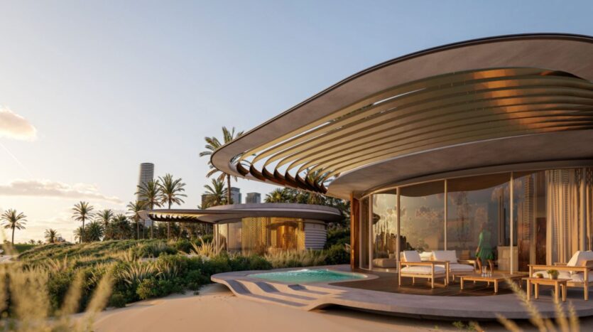A modern seaside villa at The Edition with curved architecture, large bay windows, a swimming pool, and outdoor seating surrounded by palm trees and greenery under clear skies. Another similar villa can be seen in the background.