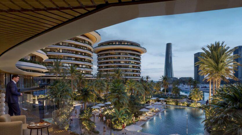 A man stands on a balcony at The Edition, overlooking a tropical complex with palm trees, a swimming pool, lounge chairs, and modern round buildings, with a marina and residential towers in the background at dusk.