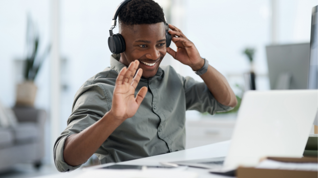 A smiling person wearing headphones sits at a desk, waving a laptop screen during a video call in a modern, bright office—discussing whether buying real estate in Dubai remotely is really possible.