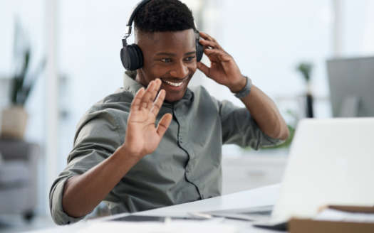 Une personne souriante portant des écouteurs est assise à un bureau, agitant l'écran d'un ordinateur portable lors d'un appel vidéo dans un bureau moderne et lumineux - discutant si acheter un bien immobilier à Dubaï à distance est vraiment possible.
