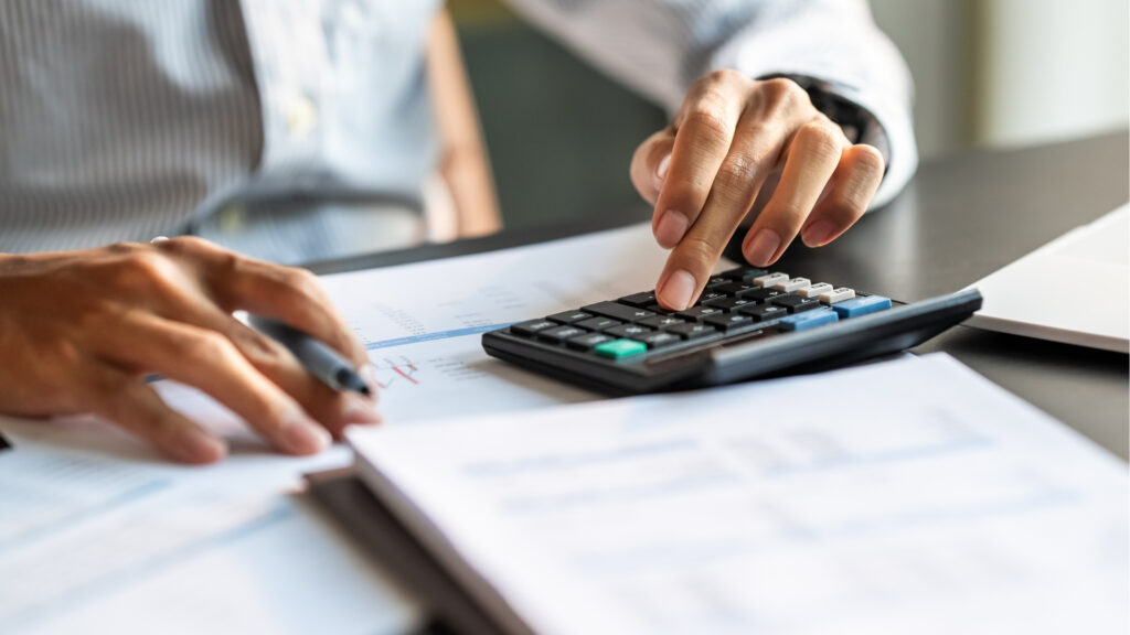A person in a striped shirt uses a calculator and pen while reviewing financial documents on a desk, figuring out how to calculate a true real estate ROI in Dubai.