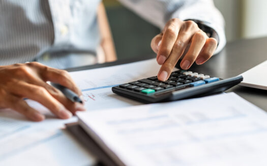 Une personne en chemise rayée utilise une calculatrice et un stylo tout en examinant des documents financiers sur un bureau, cherchant comment calculer un vrai ROI immobilier à Dubaï.